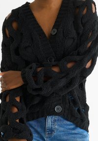 Black knitted cardigan with a textured, open weave design, featuring three large buttons and long sleeves. Worn over a black top.