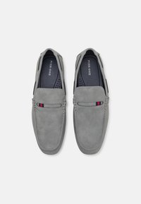 Mocassins en daim gris avec une texture lisse, présentant un détail de couture décoratif et une petite bande d'accent rouge et bleue.