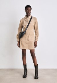 Beiges Cord-Top-Shirtkleid mit Kragen, vorderen Knöpfen und zwei Taschen, kombiniert mit knielangen schwarzen Lederstiefeln und einer schwarzen Umhängetasche.