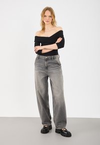 Haut noir dénudé sur les épaules associé à un jean gris taille haute et large. Le mannequin porte des chaussures à plateforme noires et épaisses. Fond uni.