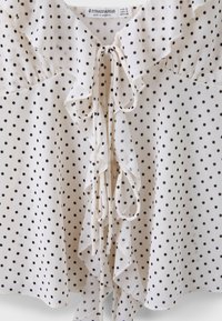 Blouse blanche à pois noirs, col à volants, détails noués à l'avant et étiquette indiquant la marque Stradivarius et la taille.
