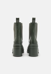 Botas de tobillo de charol verde con paneles elásticos laterales, punta redondeada y tacón grueso. Incluye dos tiradores en el tacón para facilitar su uso.