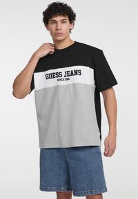 Juoda, balta ir pilka spalvų blokais išpjaustyta T marškiniai su "GUESS JEANS" užrašu. Trumpomis rankovėmis ir laisvu kirpimu. Dėvima su džinsiniais šortais.