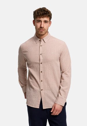 Mężczyzna w beżowej teksturowanej koszuli na długi rękaw z kołnierzykiem, stojący na jasnym, jednolitym tle.