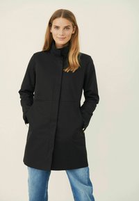 Black waterproof coat with a high collar, front pockets, and a straight silhouette. Styled with blue jeans. Smooth texture, minimal design.