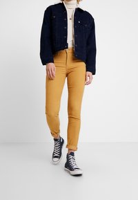 Chaqueta de mezclilla en azul marino con cierre de botones, combinada con pantalones skinny de color mostaza. El calzado presenta zapatillas altas oscuras con acentos blancos.