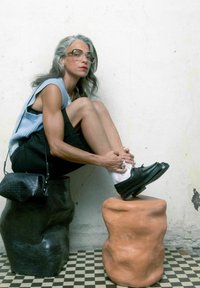 Černé a oranžové sochařské stoličky s nepravidelnými tvary podpírají osobu, která si upravuje černou botu. Viditelná texturovaná zeď a kostkovaná podlaha.