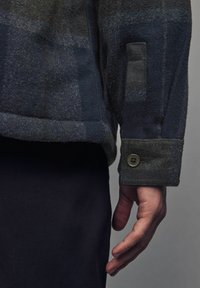 Vlnené sako s tmavomodrým károvaným vzorom, s manžetou na gombík a jemnou textúrou. Detail rukáva a ruky na tmavom pozadí.