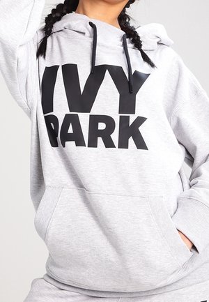 Personne portant un sweat à capuche gris clair avec un texte noir "IVY PARK", des cordons noirs et les mains partiellement dans la poche avant.