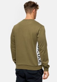Sweatshirt vert olive, en mélange de coton, col rond, poignets et ourlet côtelés, présente un panneau latéral blanc avec le texte noir "CODE." Design simple et décontracté.