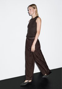Brązowy bezrękawnik zestawiony z luźnymi brązowymi spodniami i szarymi balerinami. Modelka idzie na białym tle z czarną podłogą.