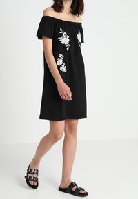 Czarna sukienka bez ramiączek z białym haftem kwiatowym. Krótkie rękawy, luźny krój i długość do kolan. Noszona z czarnymi sandałami z klamrami.
