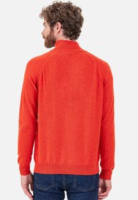 Roter Strickpullover mit hohem Kragen, Raglanärmeln und gerippten Bündchen. Weiche Textur und figurbetonte Silhouette, kombiniert mit dunkelblauen Jeans.