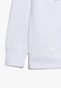 Manica e tasca di una felpa bianca con cuciture visibili e polsini a costine su uno sfondo semplice.