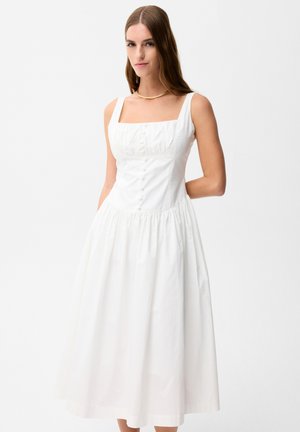 Kobieta ubrana w białą, bezrękawną midi sukienkę z guzikami z przodu i kwadratowym dekoltem, stojąca na jednolitym tle.