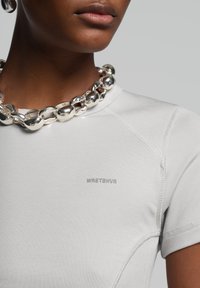 T-shirt aderente grigio chiaro con maniche corte, caratterizzato da un logo argento "WRSTBVR" e una collana in argento con catena testurizzata.