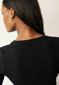 Black ribbed fabric top with a rounded neckline and a center seam detail, worn on a model showing long, straight hair and gold earrings.