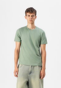 T-shirt verde din bumbac, cu mâneci scurte, guler rotund, croială lejeră. Model îmbrăcat cu pantaloni din denim de culoare albastru deschis. Material simplu, fără modele sau accente.