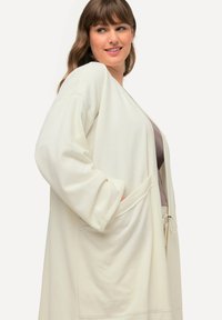 Cardigan lungo aperto in morbido tessuto crema con maniche ampie e tasche frontali. Presenta una finitura testurizzata e una vestibilità rilassata.