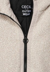 Chaqueta de forro polar beige claro con superficie texturizada, cremallera negra y una etiqueta de marca. Cuenta con capucha y un forro interior de contraste.