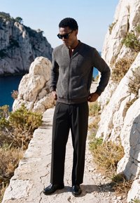 Homme portant des lunettes de soleil, une veste grise zippée, un pantalon noir et des chaussures noires se tient sur un chemin rocheux près des falaises au bord de la mer bleue sous un ciel dégagé.
