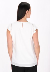 Valkoinen blouse, jossa on pitsikoristeita röyhelöillä varustetuissa hihoissa, takana avainaukko ja sileä tekstuuri. Suunniteltu rentoa istuvuutta varten.