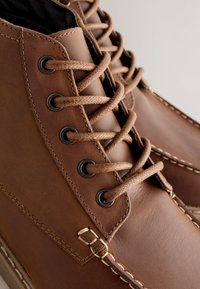 Bottes en cuir marron avec un col matelassé, des œillets noirs et des lacets marron. La semelle présente des coutures contrastantes le long des bords.