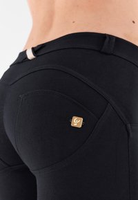 Musta leggingsit joustavasta kankaasta, joissa koristeellinen tasku kultaembleemilla ja pieni valkoinen vyötärönauhalenkki.