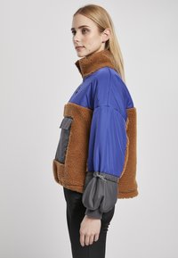 Värviline jakk, mille pealsel on sinine nailon ja allosas pruun fleece. Hõlmab halli varrukad elastsete kinnitustega ja esikoti.
