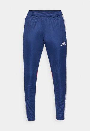 Marineblå treningsbukser laget av glatt materiale med hvite sidestriper, elastisk midjebånd og en rød detalj på den nedre kanten.