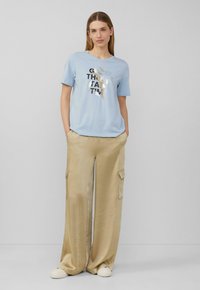 Lichtblauw T-shirt met korte mouwen met zwarte en gouden tekst "GOEDE DINGEN VERGEN TIJD", gecombineerd met beige wijde cargobroek en witte schoenen.