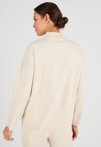 Pullover di un beige chiaro, a coste, con colletto alto e spacchi laterali. Texture morbida e vestibilità rilassata, con spalle scese.