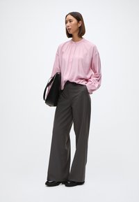 Lyserosa langermet bluse med rund hals, kombinert med vide grå bukser. Svart skulderveske og kraftige svarte sko fullfører antrekket.