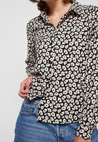 Blusa nera con un motivo floreale in crema, dotata di colletto, chiusura frontale con bottoni e maniche lunghe. Indossata con jeans blue a vita alta.