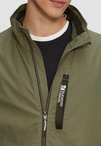 Olijfgroene jas met een hoge kraag, voormezipper en logodetail op de borst. Heeft een gladde textuur en een contrasterende binnenvoering.