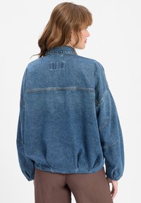 Denimjack in medium blauw met een relaxte pasvorm, voorzien van een ronde kraag, verlaagde schouders en een elastische zoom. Exposed stikseldetails.