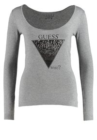Haut gris à manches longues avec un large col, présentant un motif triangle en sequins noirs et le texte "DEVINE QUI ?" en dessous. Texture de tissu douce.