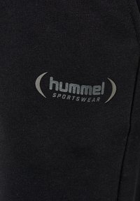 Schwarzer Sportbekleidungsstoff mit glatter Textur, auf der eine graue "hummel SPORTSWEAR"-Logoprint prominent an der Seite zu sehen ist.