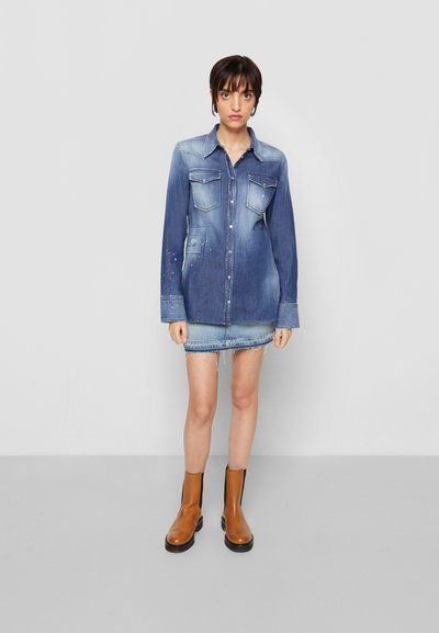 Camisa de denim de mangas longas em azul escuro, com dois bolsos no peito, ligeira descoloração e fecho de botão; usada sobre uma saia mini de denim claro e botas de tornozelo castanhas.