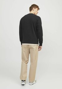 Sudadera de algodón negra con cuello redondo, combinada con pantalones de algodón beige. Modelo de pie de lado, mostrando un fit relajado y colores neutros.