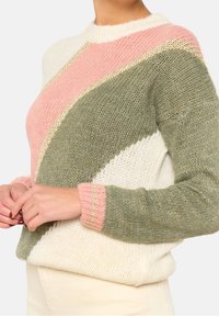 Kvinna som bär en stickad tröja med diagonala färgblock i kräm, rosa och olivgrön, som håller händerna vid midjan.