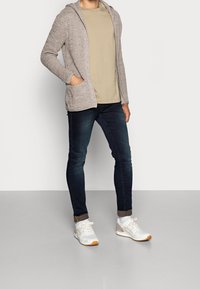 Lichtgrijze gebreide cardigan met een capuchon, beige t-shirt, donkere slim fit jeans en witte sneakers met oranje accenten. Heeft zijzakken.
