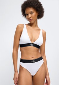 Γυναίκα που φοράει λευκό μπραλέτο Calvin Klein και εσώρουχα με ψηλή μέση και μαύρες ελαστικές ταινίες, στέκεται μπροστά σε απλό φωτεινό φόντο.