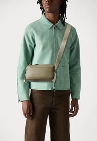 Mint green suede jacket with a zip, paired with brown trousers. A rectangular olive green crossbody bag with a strap rests on the shoulder.
