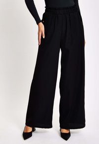 Black wide-leg pants with a ruffled elastic waistband, side pockets, and sleek fabric, paired with pointed black heels.