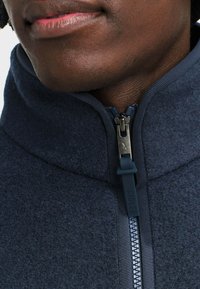 Giacca in pile blu navy con colletto alto, dotata di una cerniera metallica con un tirante in tessuto e texture a coste lungo i bordi.