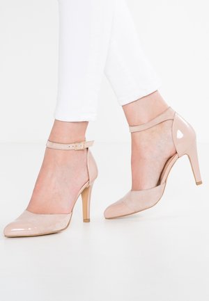Escarpins nude en cuir verni et daim à bride cheville portés avec un pantalon blanc, vue latérale des deux pieds sur une surface blanche.