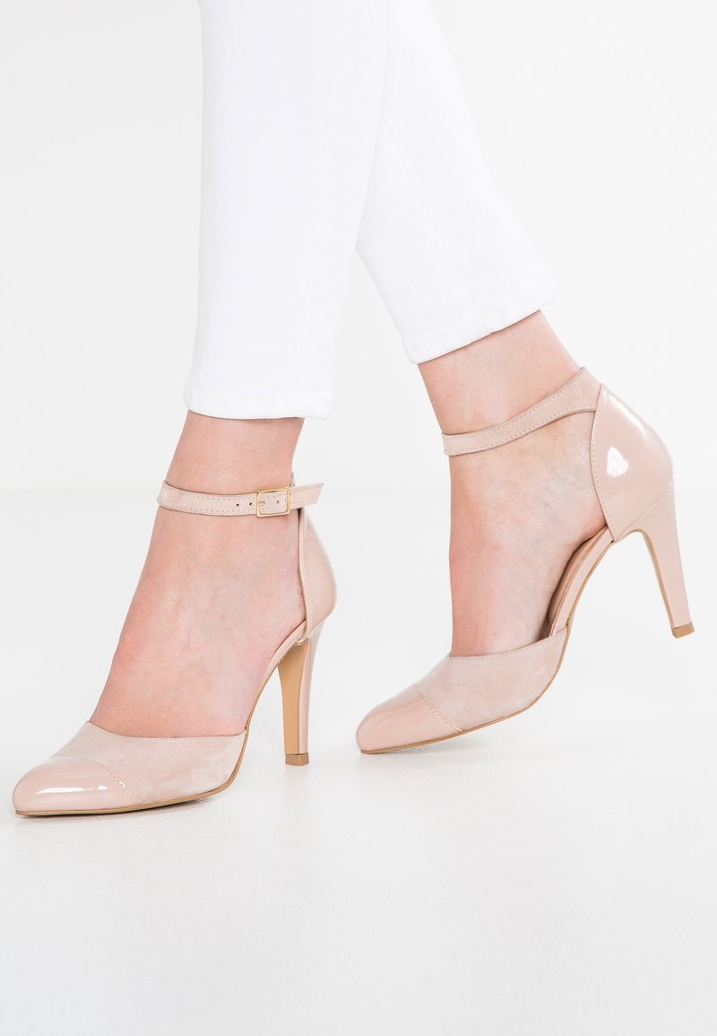 Escarpins nude en cuir verni et daim à bride cheville portés avec un pantalon blanc, vue latérale des deux pieds sur une surface blanche.