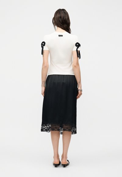 Γυναίκα με πλάτη, ντυμένη με άσπρο μπλουζάκι με κοντά μανίκια και μαύρες λεπτομέρειες φιόγκου, καθώς και μαύρη midi φούστα με δαντελένιο τελείωμα, συνδυασμένη με μαύρα τακούνια.