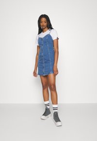 Denim dress with button-down front, adjustable straps, and blue color. Paired with a white T-shirt and gray high-top sneakers.